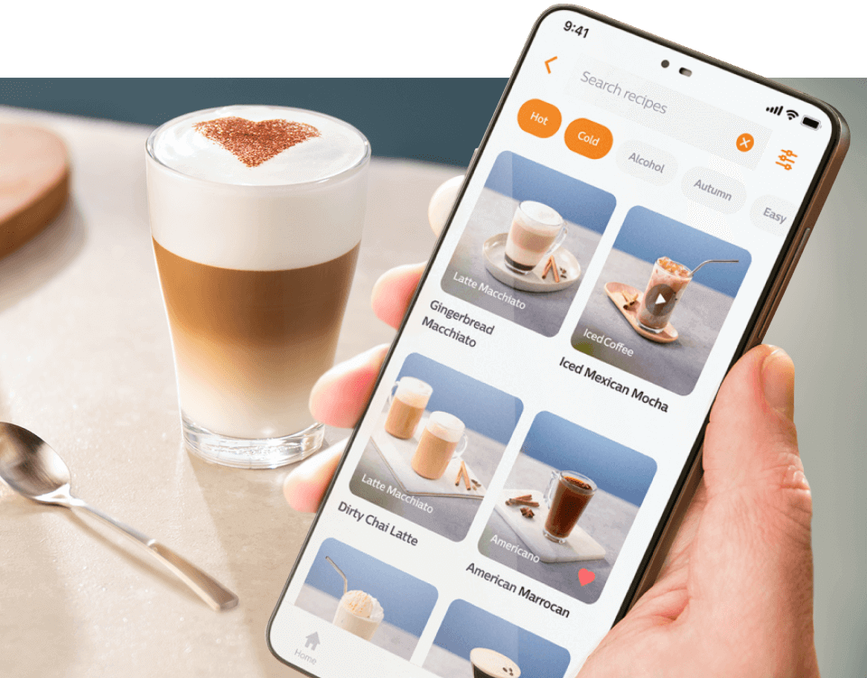 Osoba trzyma smartfon z aplikacją do przepisów na kawę, na ekranie widoczne są różne rodzaje kaw, takie jak \'Latte Macchiato\' i \'Iced Mexican Mocha\'. Obok stoi szklanka z kawą z pianką, na której wierzchu znajduje się wzór serca z kakao.