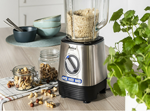 Na blacie kuchennym stoi blender kielichowy marki Amica z wypełnionym kielichem, obok znajdują się miski, słoiki z orzechami i roślina doniczkowa.