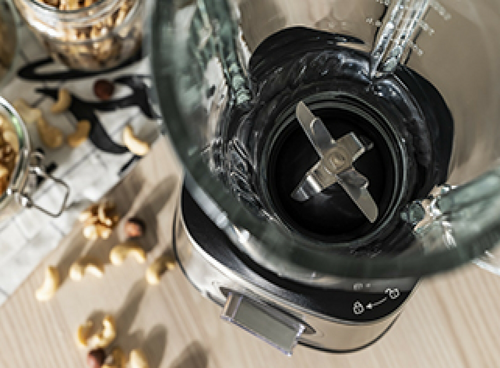 Zdjęcie przedstawia blender kielichowy z widocznymi ostrzami wewnątrz szklanego pojemnika. W tle znajdują się orzechy i inne składniki na drewnianym blacie.
