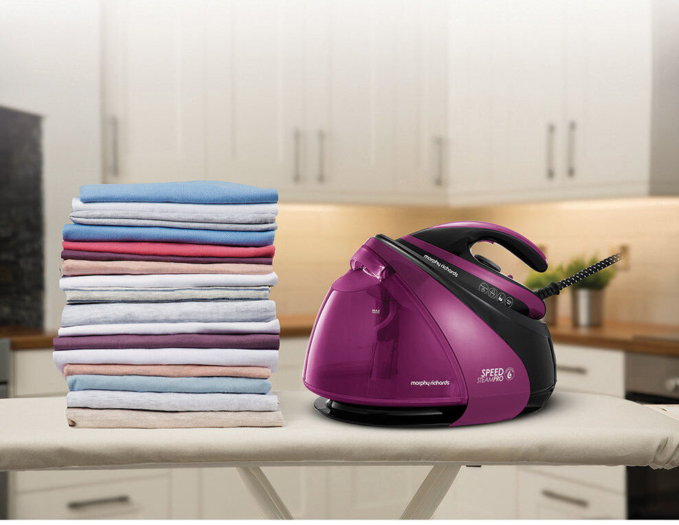 Na desce do prasowania znajduje się generator pary w kolorze fioletowym z napisem \'morphy richards SPEED STEAM PRO\'. Obok leży stos kolorowych, starannie złożonych ubrań.