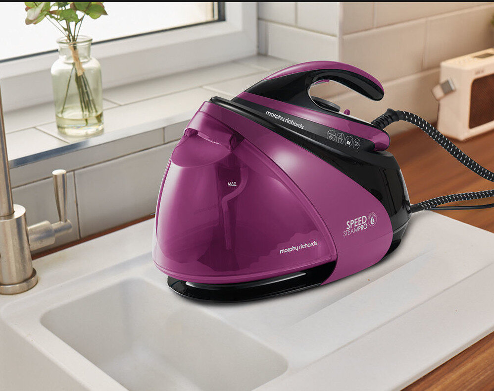 Na obrazie znajduje się fioletowy generator pary z napisem \'morphy richards\' i \'SPEED STEAM PRO\'. Urządzenie stoi na blacie kuchennym obok zlewu.