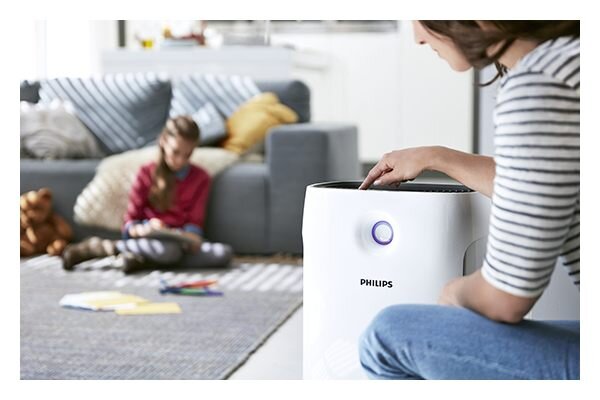 Na pierwszym planie znajduje się biały oczyszczacz powietrza z widocznym logo PHILIPS i podświetlanym przyciskiem. W tle widać osobę siedzącą na dywanie w salonie z kanapą i poduszkami.