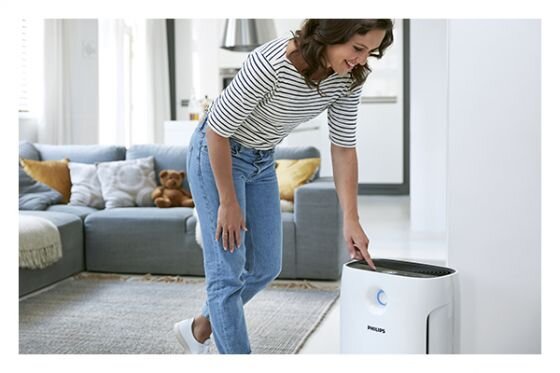Osoba obsługuje oczyszczacz powietrza marki PHILIPS w jasnym, nowoczesnym salonie. W tle widoczna jest szara sofa z poduszkami i pluszowym misiem.
