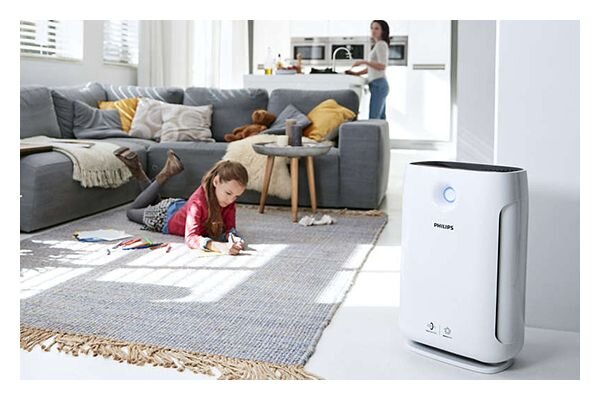 W jasnym salonie na dywanie leży dziecko rysujące kredkami, obok stoi oczyszczacz powietrza z logo PHILIPS. W tle widać sofę z poduszkami i osobę w kuchni.