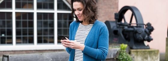 Osoba stoi na zewnątrz, trzymając w ręku smartfon. W tle widoczna jest duża maszyna przemysłowa i budynek z oknami.