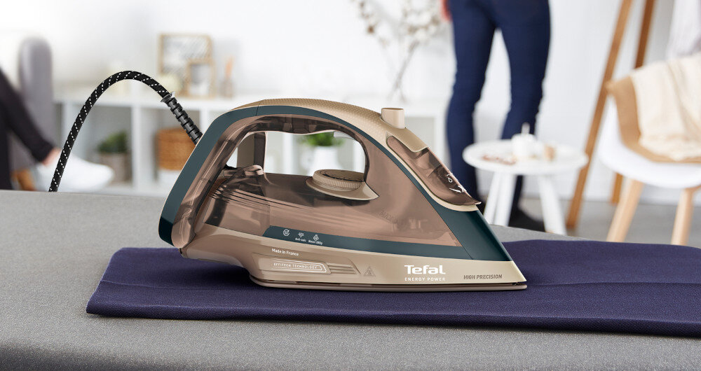 Żelazko w kolorze brązowym z napisem \'Tefal Energy Power\' stoi na desce do prasowania. W tle widoczne są meble i osoba w pomieszczeniu.
