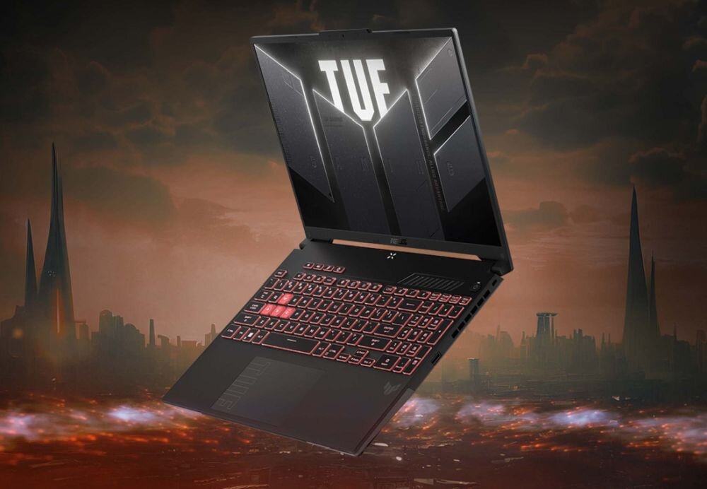 Laptop z podświetlaną klawiaturą i otwartym ekranem, na którym widnieje napis \'TUF\'. W tle widoczny jest krajobraz miasta o zmierzchu z wysokimi budynkami.