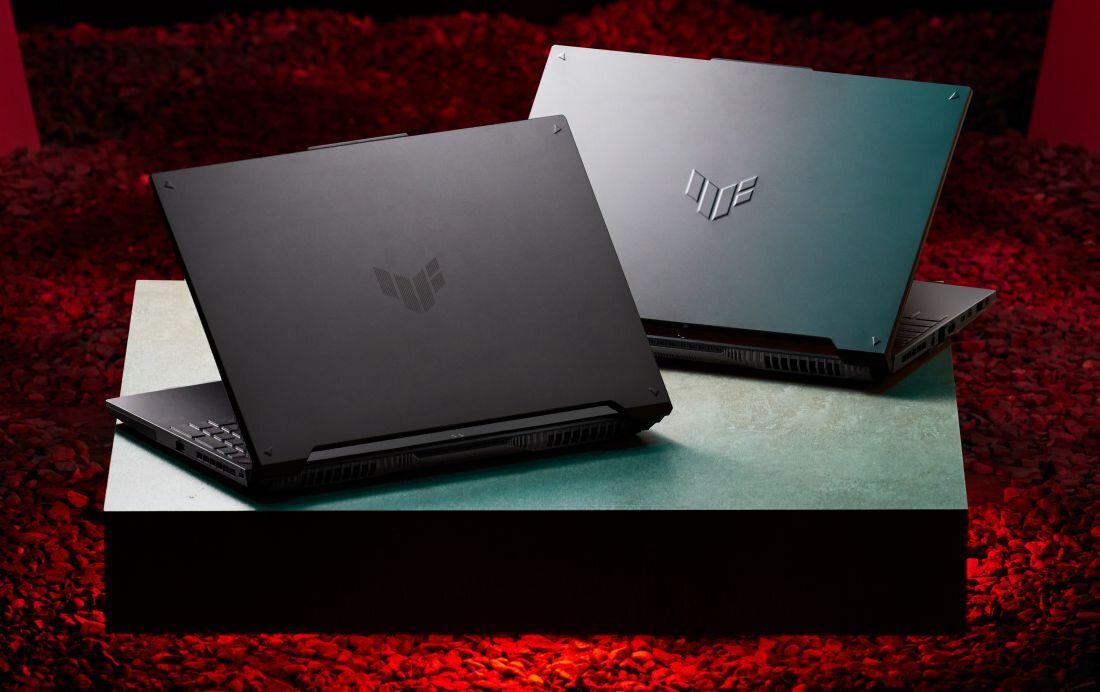 Dwa laptopy z widocznym logo TUF na pokrywach, ustawione na tle czerwonego oświetlenia. Laptopy są częściowo otwarte, ukazując klawiatury.