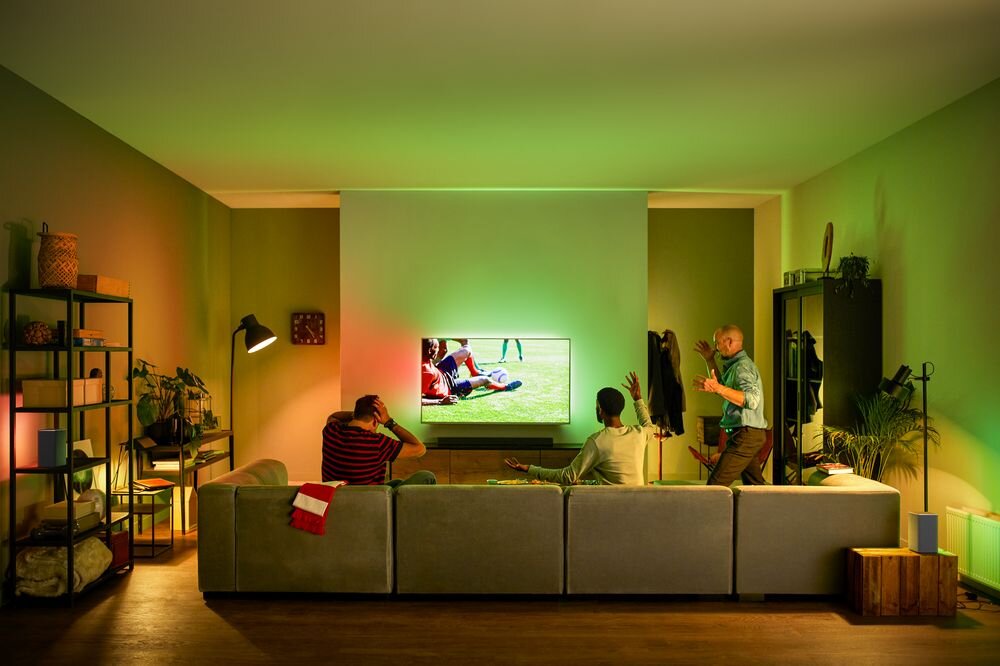 W salonie znajduje się telewizor wyświetlający obraz z zielonym oświetleniem Ambilight na ścianie. W pomieszczeniu są trzy osoby, a wnętrze jest przytulnie urządzone z roślinami i półkami.