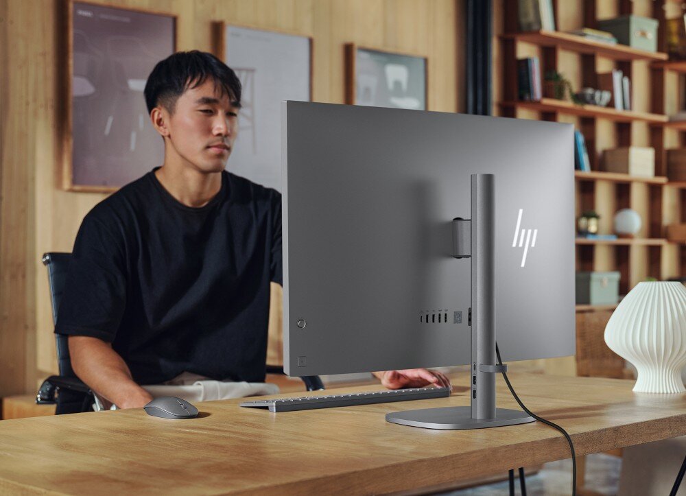 Komputer All in One HP OmniStudio X 32-C0005NW Mężczyzna siedzi przy drewnianym biurku przed tyłem obudowy monitora komputera All-in-One, na którym leży także klawiatura i mysz