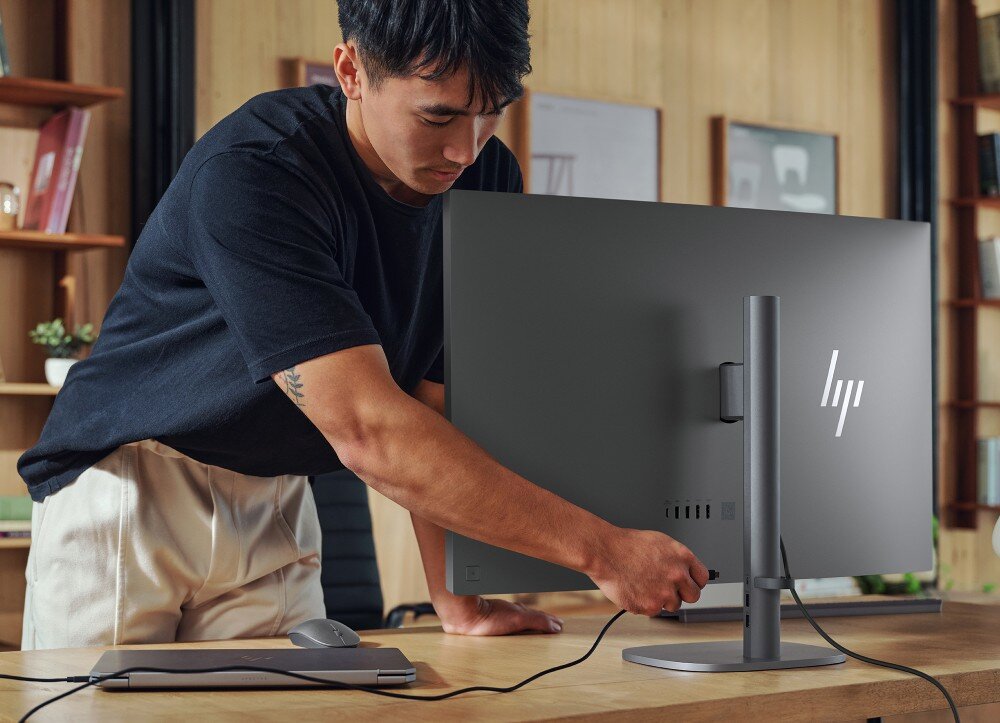 Komputer All in One HP OmniStudio X 32-C0005NW Mężczyzna pochyla się nad drewnianym biurkiem i podłącza przewód z boku tylnej obudowy monitora, do którego podpięty jest laptop
