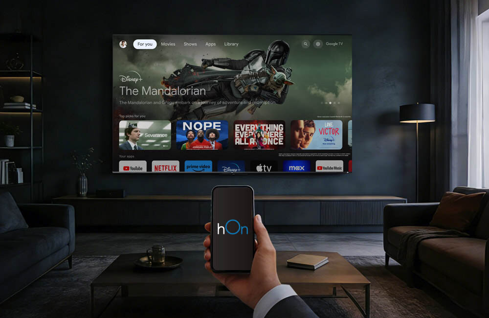 W salonie na ścianie wisi duży telewizor wyświetlający interfejs Google TV z różnymi aplikacjami i filmami, w tym \'The Mandalorian\'. Na pierwszym planie osoba trzyma smartfon z aplikacją \'hOn\' na ekranie.