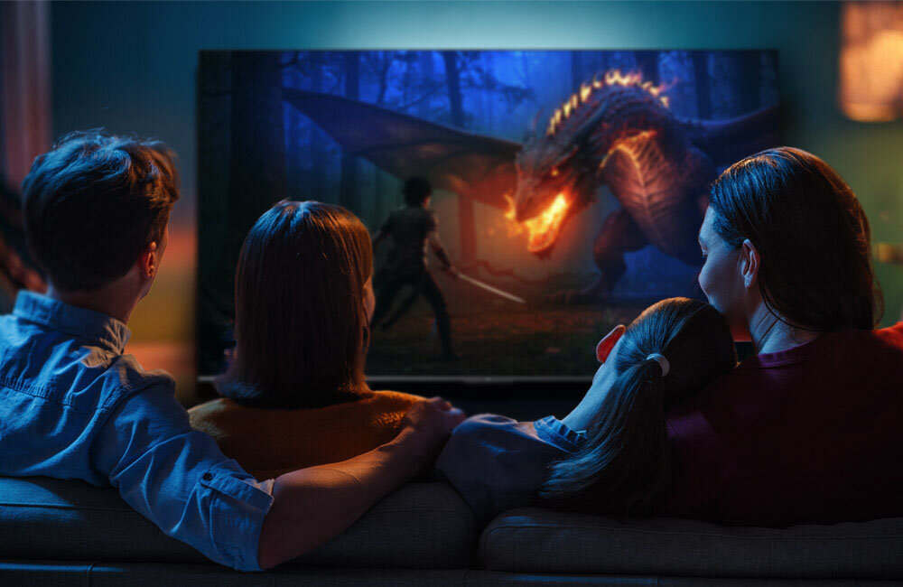 Grupa osób siedzi na kanapie i ogląda telewizor, na którym wyświetlana jest scena z filmu fantasy z postacią walczącą ze smokiem. Pomieszczenie jest przyciemnione, a ekran telewizora jest głównym źródłem światła.