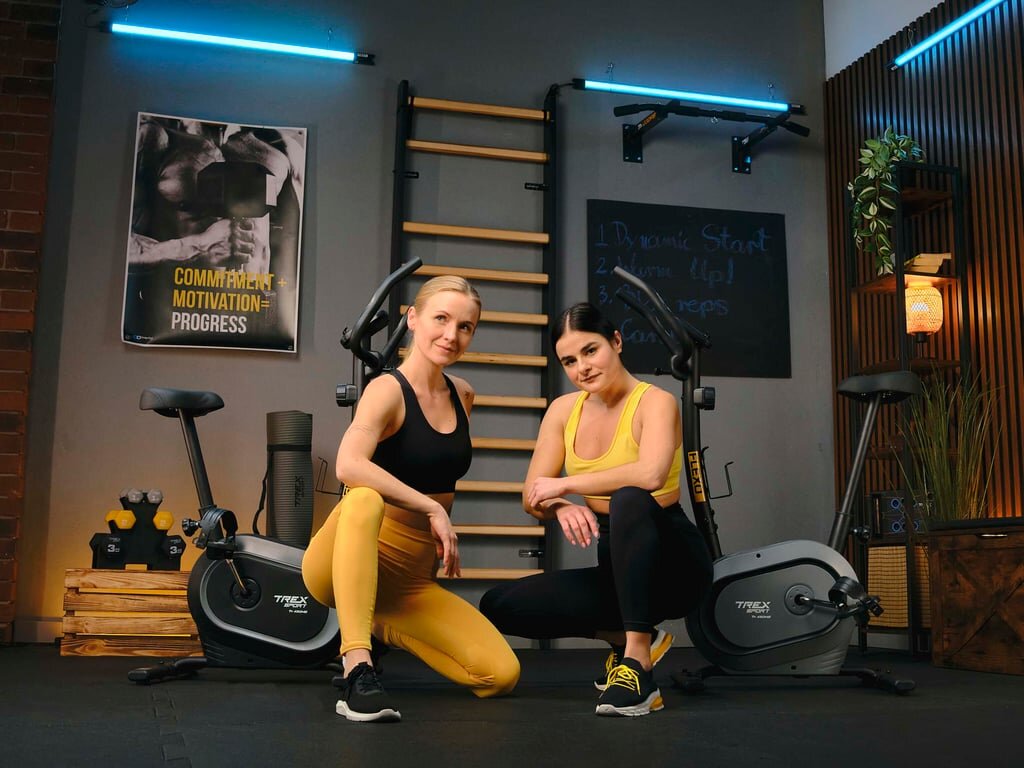 Dwie osoby w sportowych strojach siedzą obok dwóch rowerów stacjonarnych w pomieszczeniu do ćwiczeń. Na ścianie widoczny jest plakat z napisem \'COMMITMENT + MOTIVATION = PROGRESS\'.