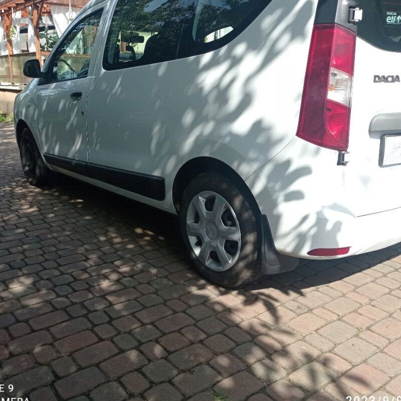 Biały samochód zaparkowany na brukowanej nawierzchni, z widocznym logo \'Dacia\' na tylnej części. Na tylnym kole zamontowany jest chlapacz.