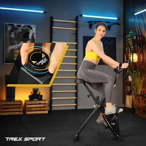 Osoba ćwiczy na czarnym rowerze stacjonarnym w pomieszczeniu z drewnianymi elementami i plakatem na ścianie. W lewym dolnym rogu widoczne jest logo \'TREX SPORT\'.