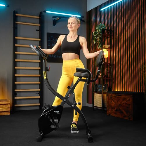 Kobieta w sportowym stroju stoi obok czarnego rowerka stacjonarnego magnetycznego Xbike PYRO TX-360XB w pomieszczeniu z drewnianymi elementami dekoracyjnymi. Na rowerku widoczne jest logo \'PYRO\'.