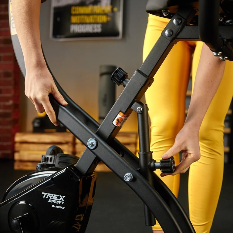 Osoba reguluje mechanizm rowerka stacjonarnego, na którym widoczne jest logo \'TREX SPORT\'. W tle znajdują się drewniane skrzynki i plakat na ścianie.