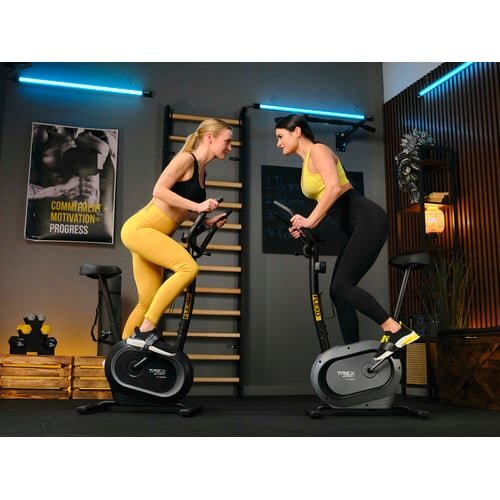 Dwie osoby ćwiczą na rowerach stacjonarnych w pomieszczeniu z nowoczesnym wystrojem. Na ścianie widoczny jest plakat z napisem \'COMMITMENT MOTIVATION PROGRESS\'.