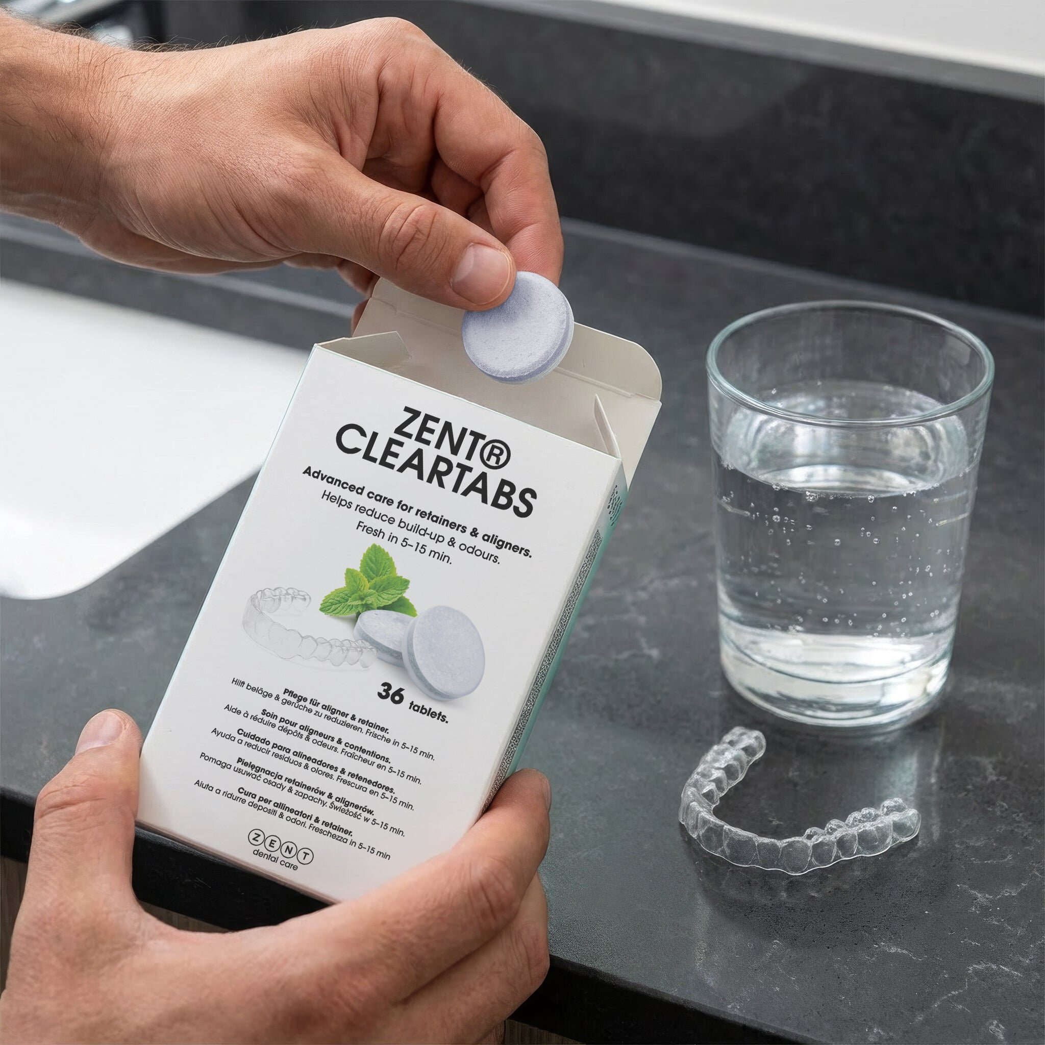 Osoba trzyma opakowanie tabletek czyszczących ZENT CLEARTABS, a obok znajduje się szklanka z wodą i przezroczysta nakładka na zęby. Na opakowaniu widoczny jest tekst: \'ZENT CLEARTABS Advanced care for retainers & aligners. Helps reduce build-up & odours. Fresh in 5-15 min. 36 tablets.\'