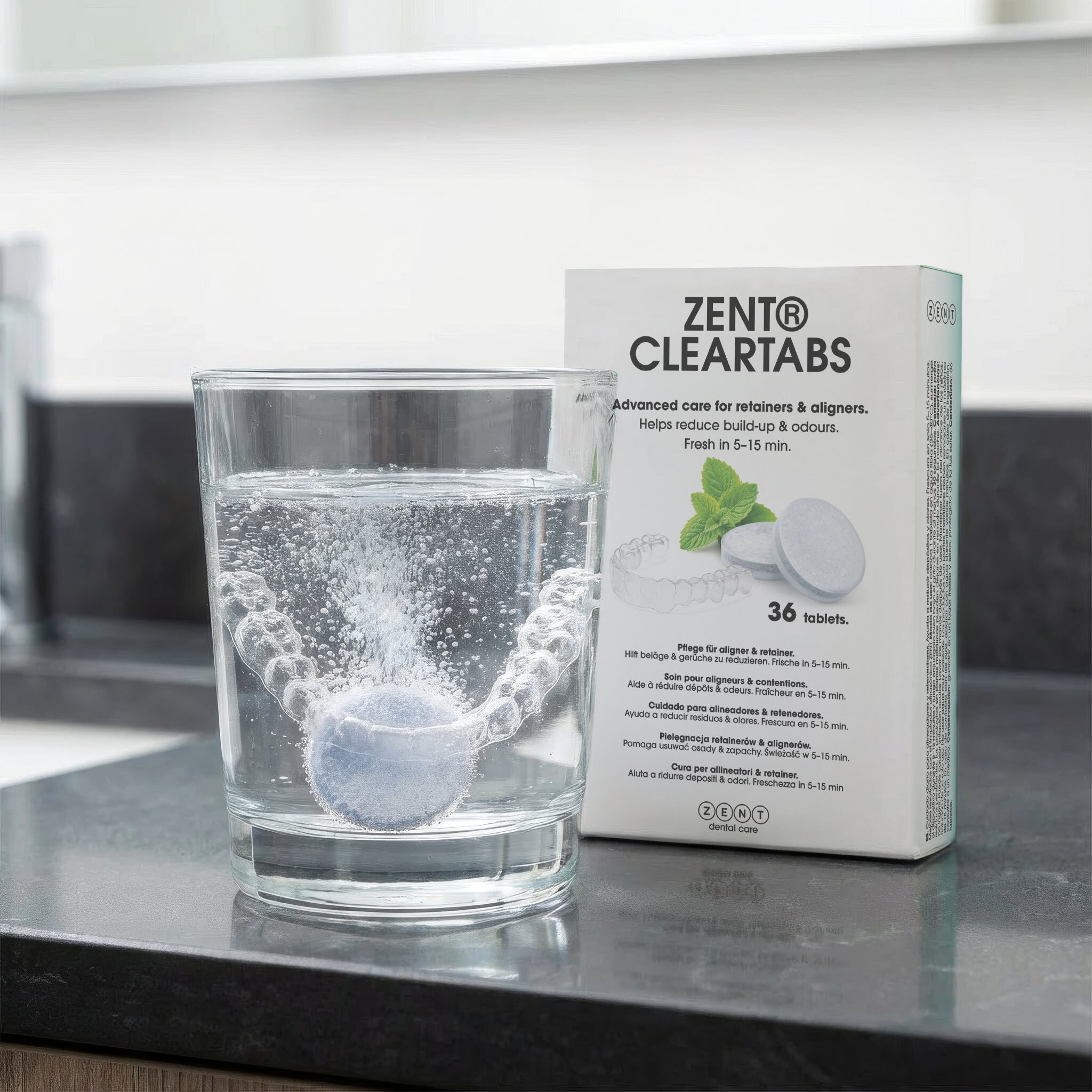 Na obrazie widać szklankę z wodą, w której rozpuszcza się tabletka czyszcząca, oraz opakowanie produktu ZENT Cleartabs z napisem \'Advanced care for retainers & aligners. Helps reduce build-up & odours. Fresh in 5-15 min. 36 tablets.\'.