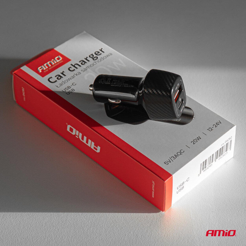 Na obrazie widoczna jest czarna ładowarka samochodowa z dwoma portami USB, umieszczona na czerwono-białym opakowaniu z napisem \'Car charger Ładowarka samochodowa\'. Logo \'AMiO\' znajduje się na opakowaniu i ładowarce.