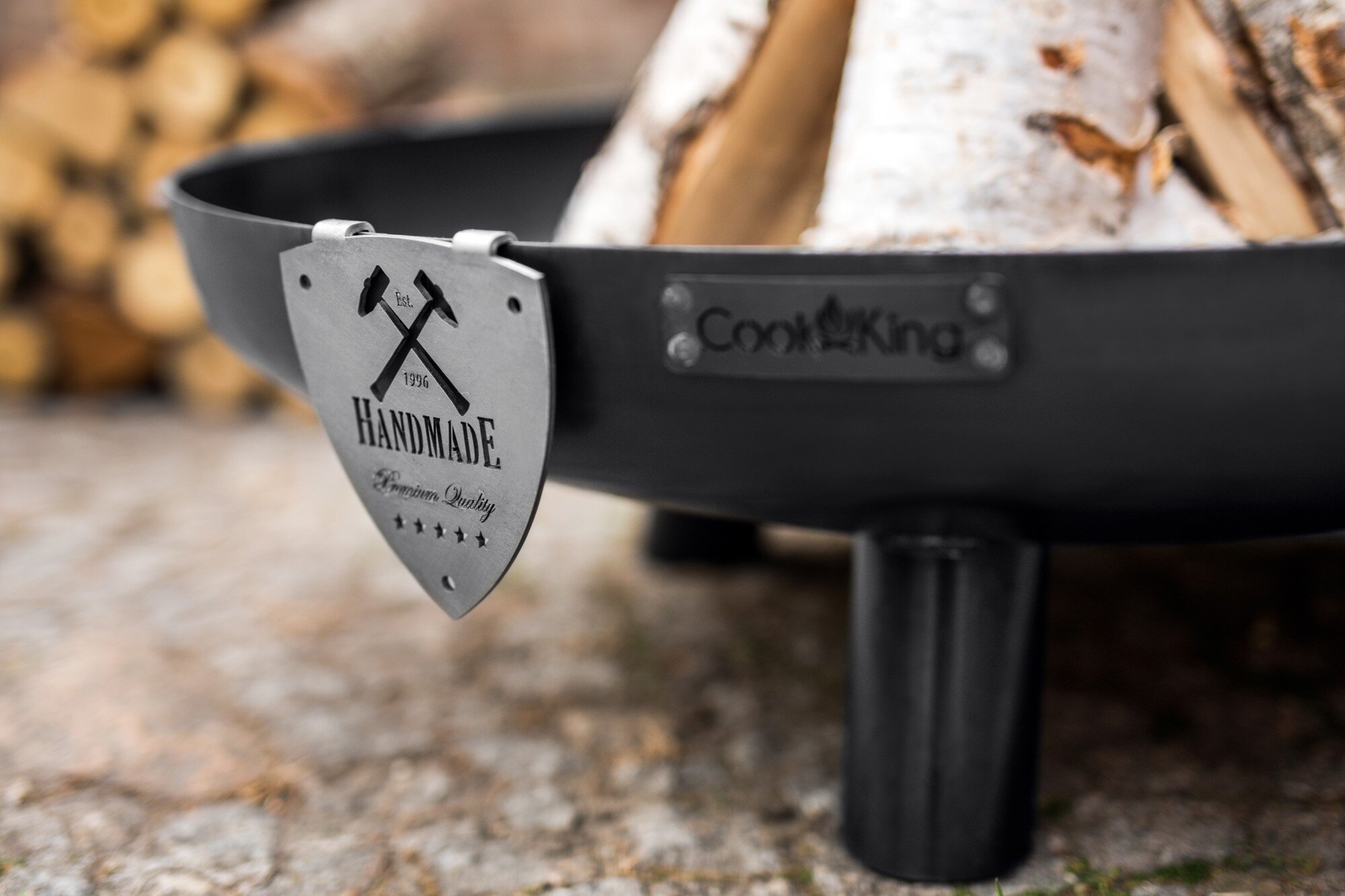 Czarne palenisko ogrodowe z metalową tabliczką z napisem \'HANDMADE Premium Quality\' oraz logo \'Cook King\'. Wewnątrz paleniska znajdują się ułożone kawałki drewna.