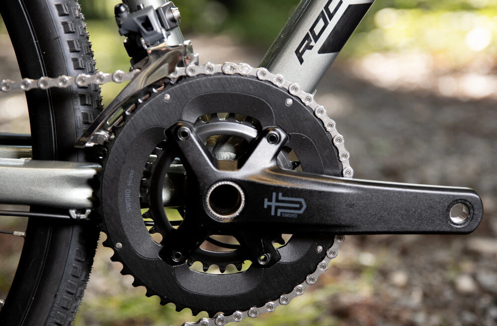 Rower gravel ROCK MACHINE Lukk 40 U19 28 cali Zielony rozmazane tło zbliżenie na korbę wkład suportu SHIMANO BSA korba PROWHEEL FC-G7000-46 kaseta MicroSHIFT CS-H105 o zakresie 11–38T odpowiednie przełożenie szeroki zakres kasety odpowiednio dobrane komponenty