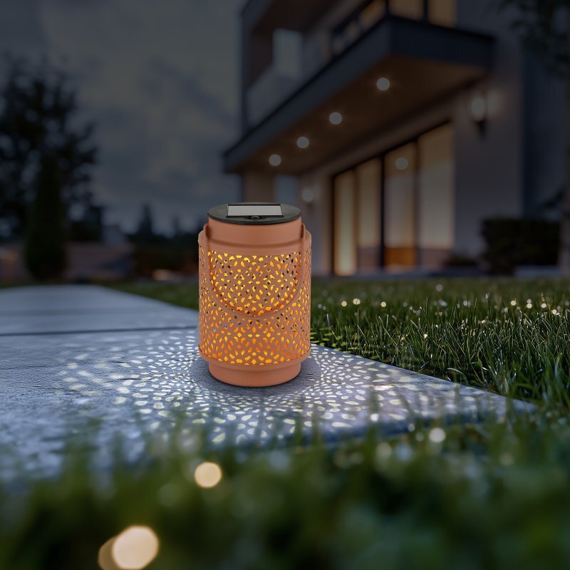 Na obrazie widoczna jest przenośna lampa solarna LED o ażurowym wzorze, emitująca ciepłe światło na chodnik i trawę. W tle znajduje się nowoczesny budynek z oświetlonymi oknami.