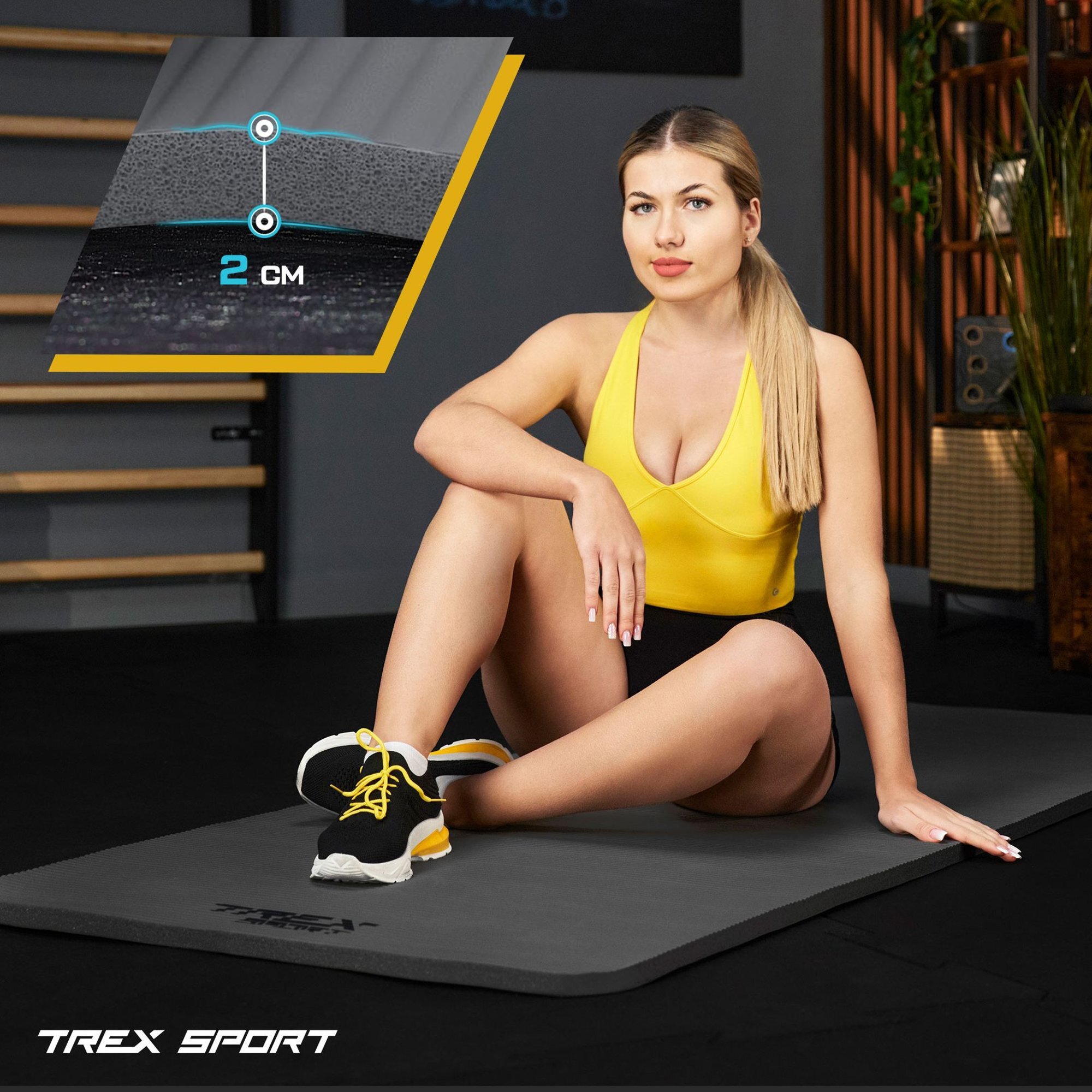 Kobieta w żółtym stroju sportowym siedzi na szarej macie do ćwiczeń. W lewym górnym rogu widoczna jest grafika przedstawiająca przekrój maty z napisem \'2 CM\'.