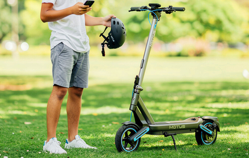 Osoba stoi na trawie obok elektrycznej hulajnogi MOTUS Scooty, trzymając kask i telefon. Hulajnoga ma widoczne logo \'MOTUS\' na boku.
