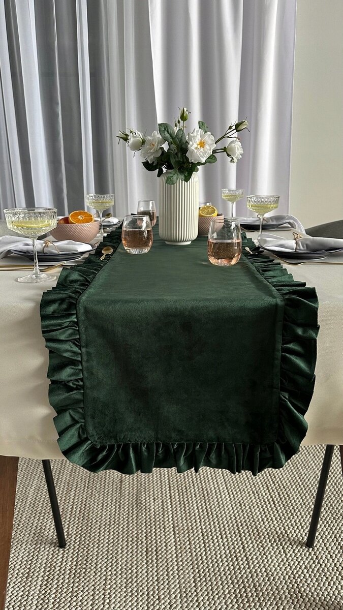 Na stole znajduje się zielony bieżnik dekoracyjny z falbankami, na którym ustawiono szklanki i kieliszki. W tle widoczny jest biały wazon z kwiatami oraz zastawa stołowa.
