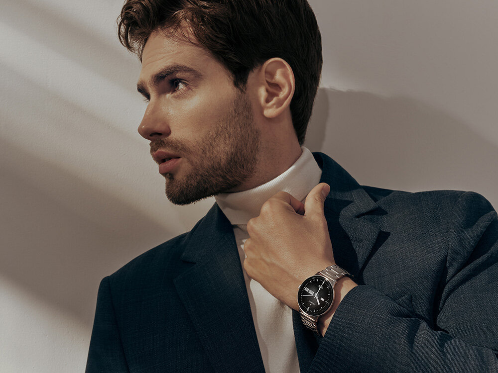 Mężczyzna w eleganckim garniturze prezentuje srebrny smartwatch na nadgarstku. Tło jest neutralne, a światło pada na twarz i zegarek.