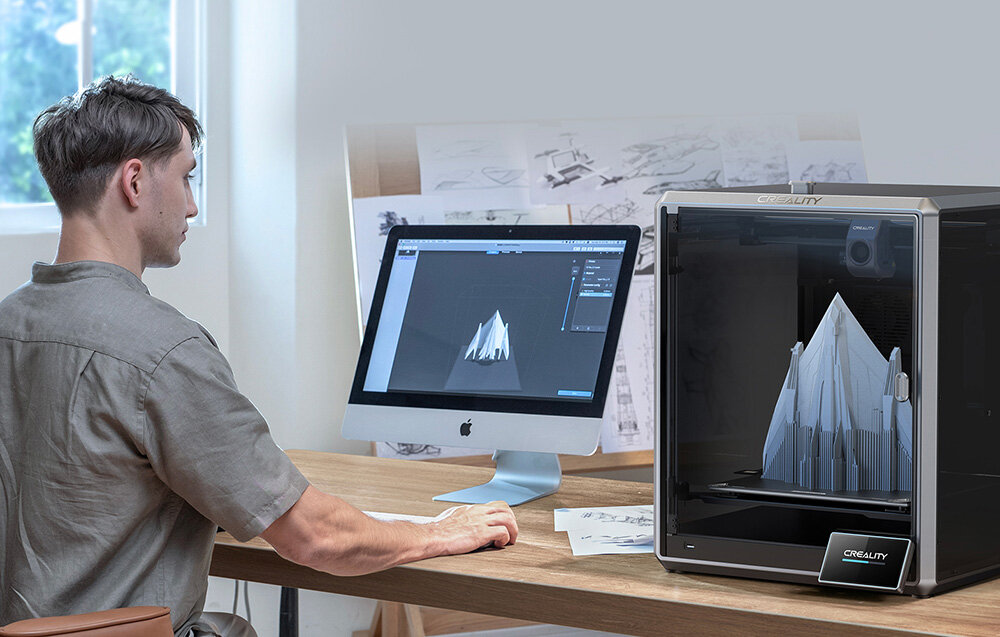 Osoba siedzi przy biurku, na którym znajduje się komputer z wyświetlonym modelem 3D oraz drukarka 3D CREALITY z wydrukowanym obiektem. W tle widoczne są szkice techniczne na tablicy.