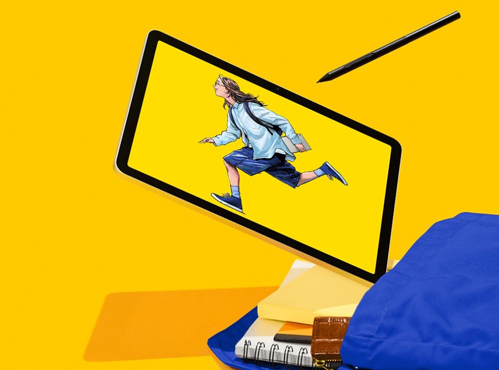 Tablet graficzny z wyświetlaczem przedstawiającym postać biegnącej osoby na żółtym tle. Obok tabletu znajduje się rysik oraz stos książek i zeszytów z niebieskim plecakiem.