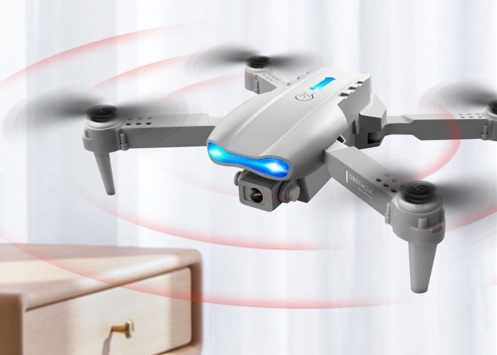 Na obrazie widoczny jest szary dron z niebieskim światłem z przodu, unoszący się w powietrzu. W tle znajduje się zasłona i fragment drewnianego mebla.