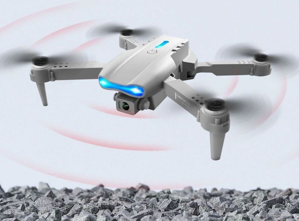 Na obrazie widoczny jest szary dron z czterema wirnikami, unoszący się nad kamienistym podłożem. Dron ma niebieskie światło z przodu i widoczny napis \'EXTRALINK\' na jednym z ramion.