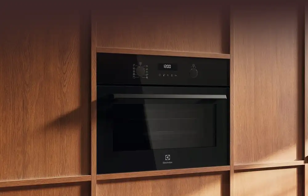 Czarna kuchenka mikrofalowa wbudowana w drewnianą zabudowę kuchenną. Na panelu widoczny jest wyświetlacz z godziną 12:00 oraz logo Electrolux.