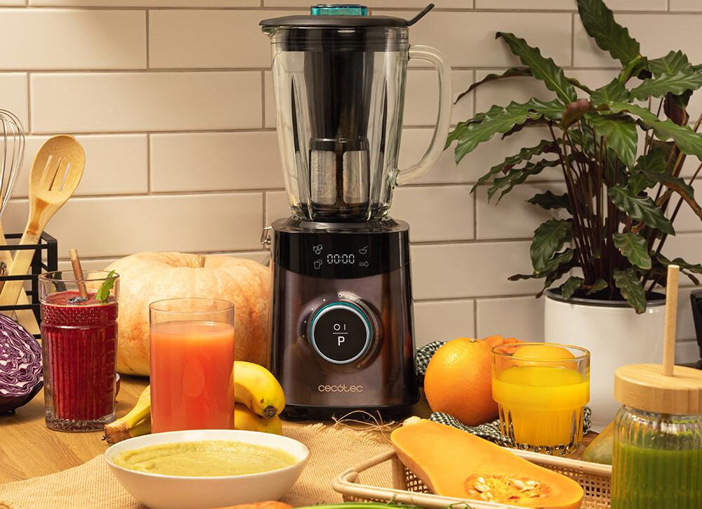 Na blacie kuchennym stoi blender kielichowy z logo \'cecotec\', otoczony różnymi owocami i napojami. W tle widoczne są białe płytki ścienne oraz roślina doniczkowa.