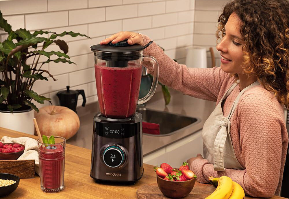 Kobieta obsługuje blender kielichowy z czerwonym napojem w kuchni. Na blacie znajdują się owoce, szklanka z napojem i logo \'cecotec\' na blenderze.
