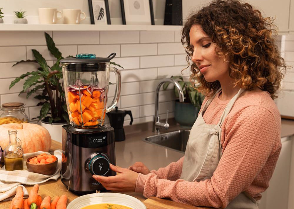 Kobieta obsługuje blender kielichowy z pokrojonymi warzywami w kuchni. Na blacie znajdują się różne składniki, a w tle widać zlew i roślinę doniczkową.