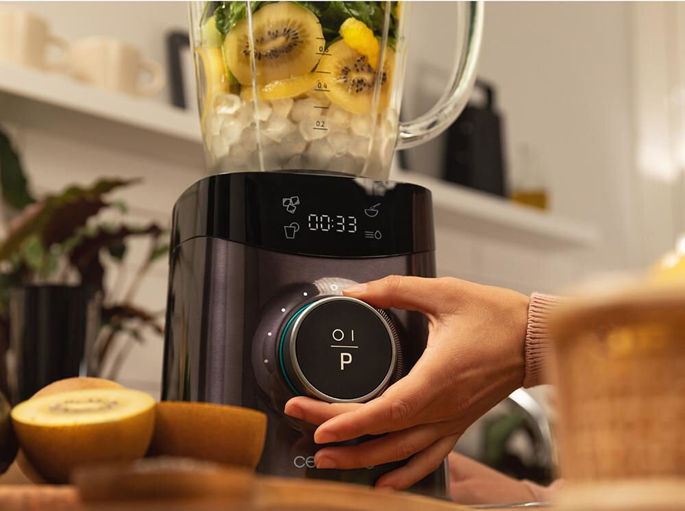 Osoba obsługuje blender kielichowy z wyświetlaczem pokazującym czas 00:33 i symbolem kubka. W kielichu znajdują się owoce i lód, a na blacie leży przekrojony owoc kiwi.