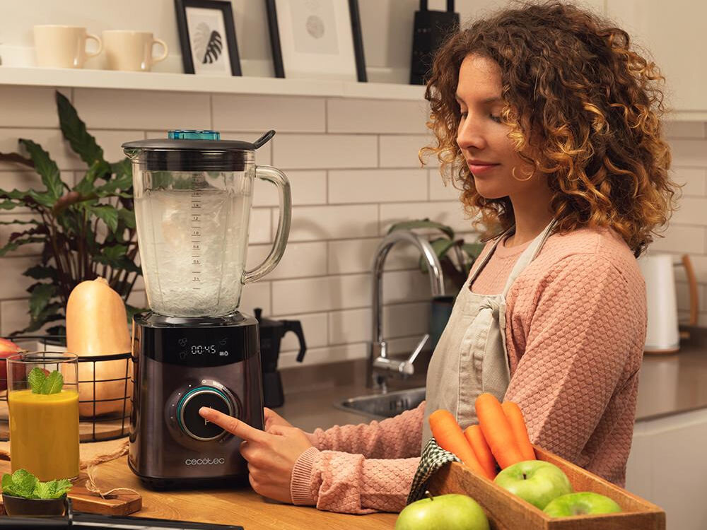 Kobieta w kuchni obsługuje blender kielichowy z wyświetlaczem pokazującym \'00:45\'. Na blacie znajdują się owoce, warzywa i szklanka z napojem.