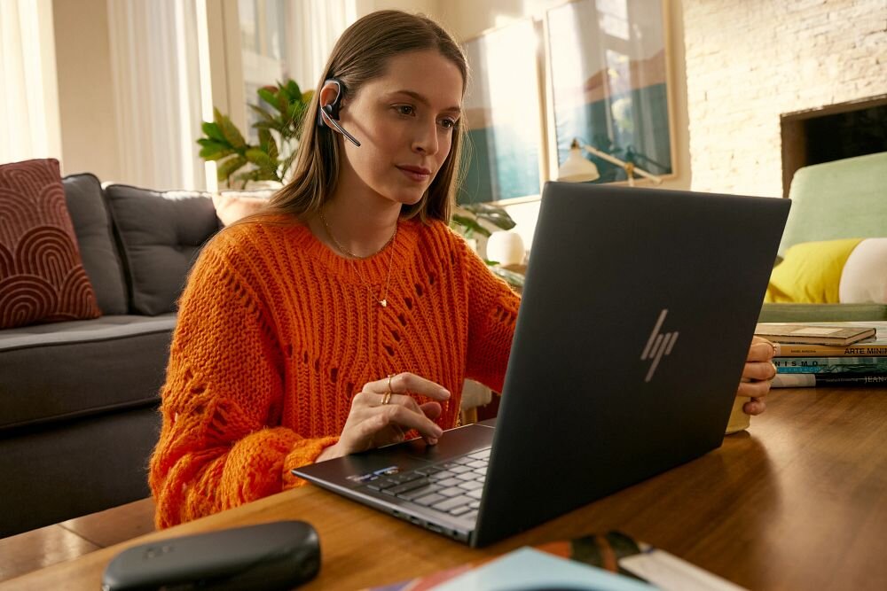 Osoba w pomarańczowym swetrze siedzi przy stole, używając laptopa z logo HP. Na głowie ma zestaw słuchawkowy z mikrofonem.