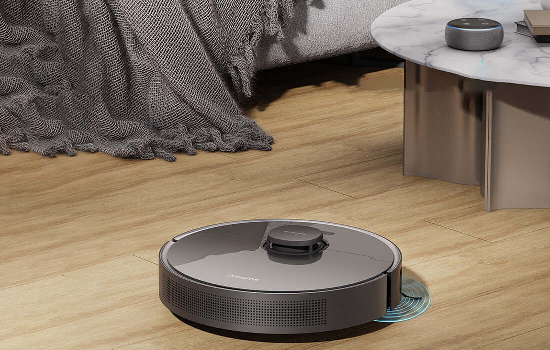 Na drewnianej podłodze znajduje się robot sprzątający z widocznym napisem \'Dreame\'. W tle widać fragment sofy z kocem oraz stolik z marmurowym blatem.