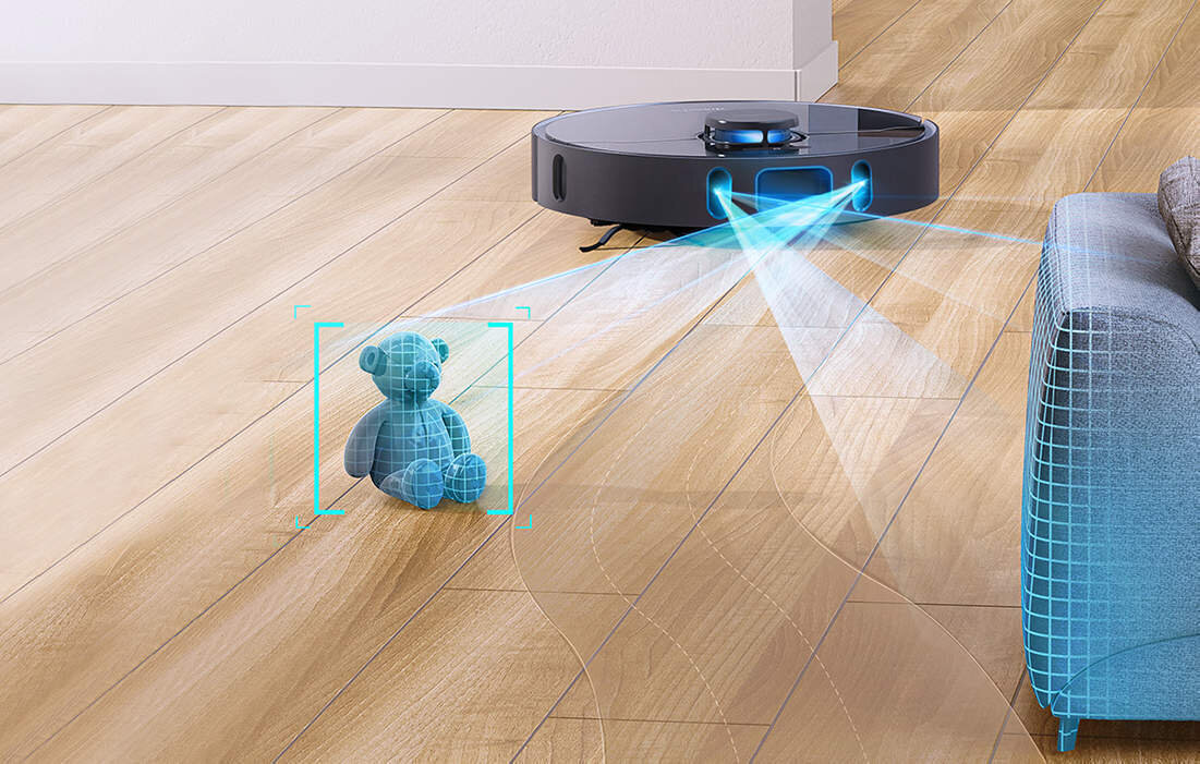 Robot sprzątający porusza się po drewnianej podłodze, emitując niebieskie światło w kierunku pluszowego misia i kanapy. Miś i kanapa są oznaczone siatką 3D, co sugeruje funkcję skanowania przeszkód.