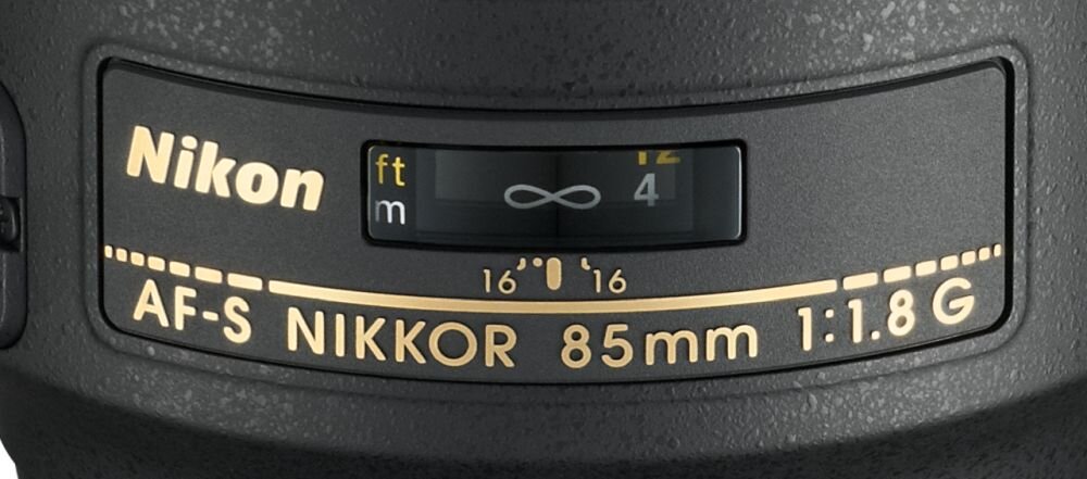 Obraz przedstawia fragment obiektywu z widocznym logo \'Nikon\' oraz oznaczeniami \'AF-S NIKKOR 85mm 1:1.8G\'.