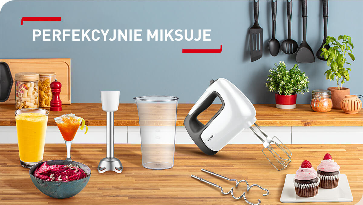 Na blacie kuchennym znajduje się mikser ręczny z różnymi akcesoriami, obok napoje i deser. Na ścianie wisi zestaw kuchennych przyborów, a nad nimi widnieje napis \'PERFEKCYJNIE MIKSUJE\'.