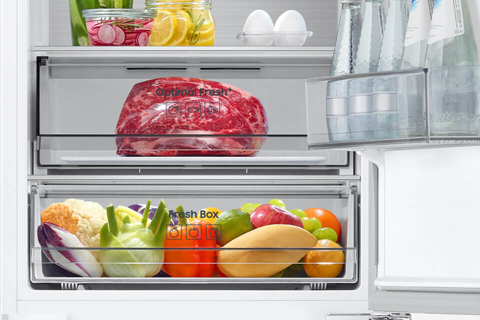 Wnętrze lodówki z widocznymi szufladami oznaczonymi jako \'Optimal Fresh\' i \'Fresh Box\', wypełnionymi mięsem oraz różnorodnymi warzywami i owocami. Na drzwiach znajdują się butelki z wodą, a na górnej półce jajka i słoiki z cytrynami oraz rzodkiewkami.