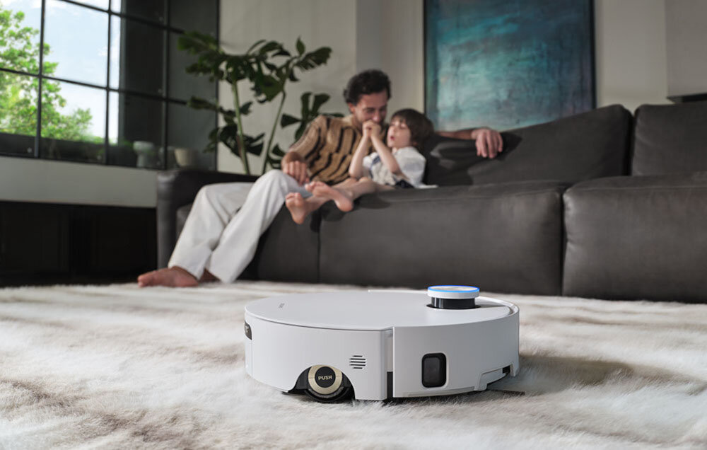 Na dywanie w salonie znajduje się biały robot sprzątający. W tle widać kanapę, na której siedzą dwie osoby.
